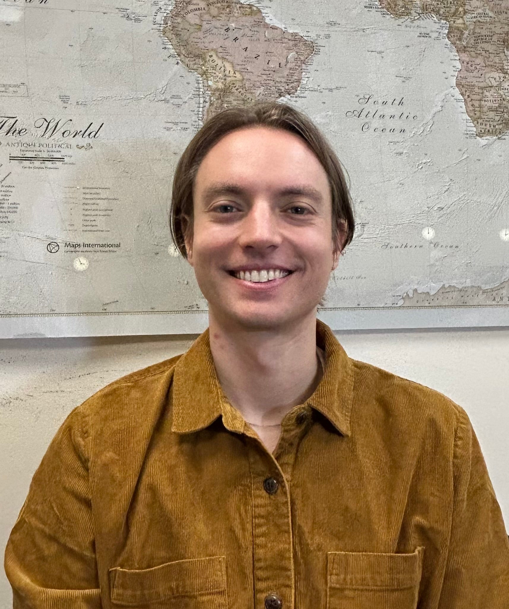 Sasha, who has fair skin and brown hair, wears a brown top while standing in front of a map