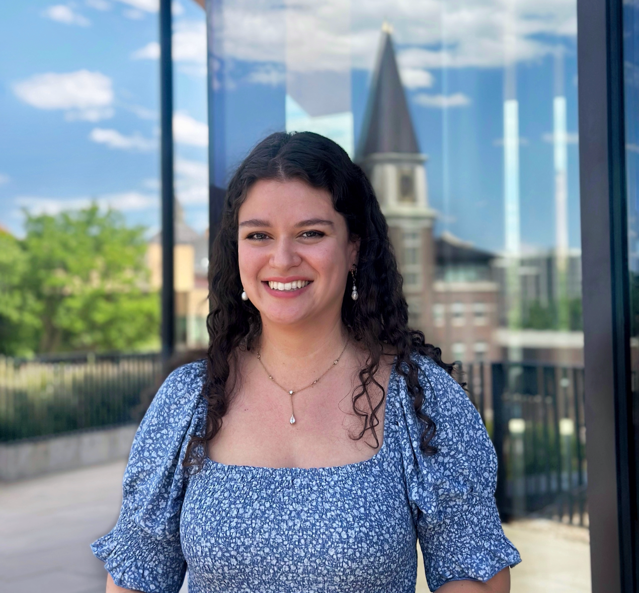 White woman smiling with brown hair, brown eyes in a blue dress, standing in front of window reflection DU buildings