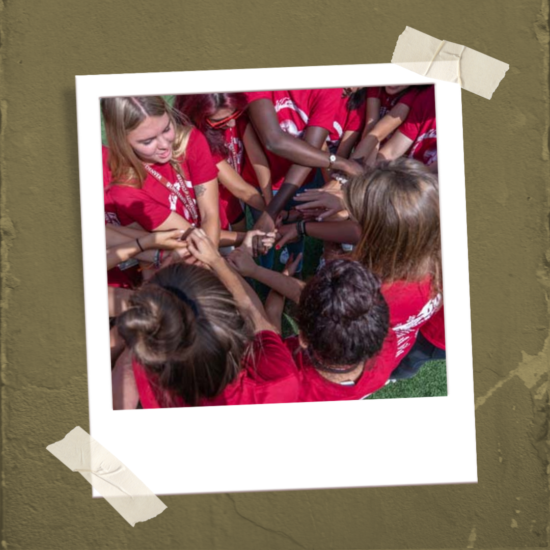 polaroid of a group of new DU students in matching red shirts with their hands in the middle of a circle for a teambuilding activity.