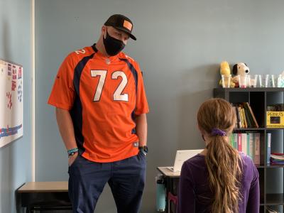 garett bolles wearing a Denver Broncos football jersey with his hands in pockets talking to a girl in a classroom