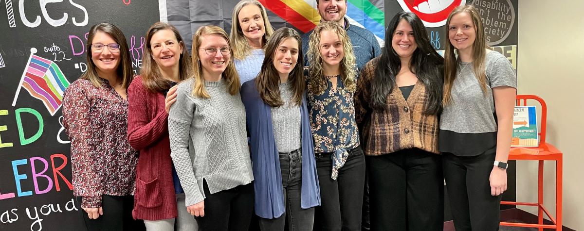 The DSP staff stands in front of a decorated chalkboard wall about about Disability Pride. The Disability Pride flag hangs in the background. 